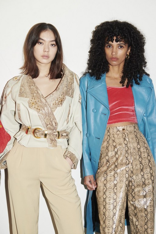 Two women in fashionable clothing stand side-by-side in a studio setting. One woman has long, dark hair and wears a snakeskin-patterned outfit.