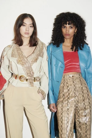 Two women in fashionable clothing stand side-by-side in a studio setting. One woman has long, dark hair and wears a snakeskin-patterned outfit.