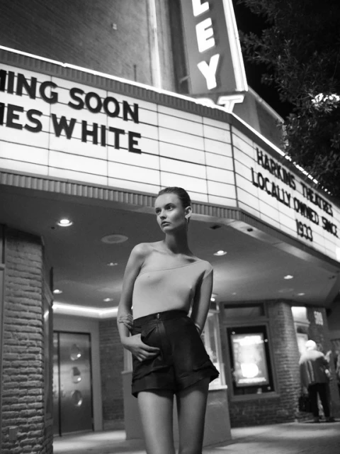 Black and white portrait of a woman in shorts standing in front of a theater at night, with illuminated marquee signage.