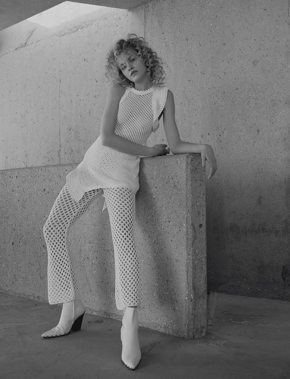 A curly-haired fashion model poses in a white mesh outfit with white boots in a concrete setting. The image is in black and white.