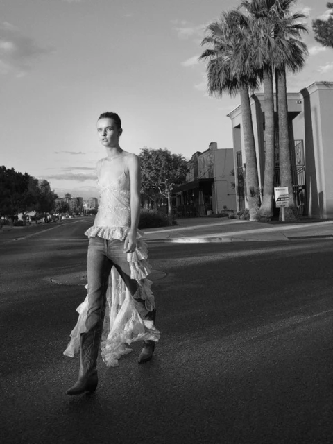 Black and white fashion shot of a woman in a dress and cowboy boots walking on a street with palm trees and buildings.