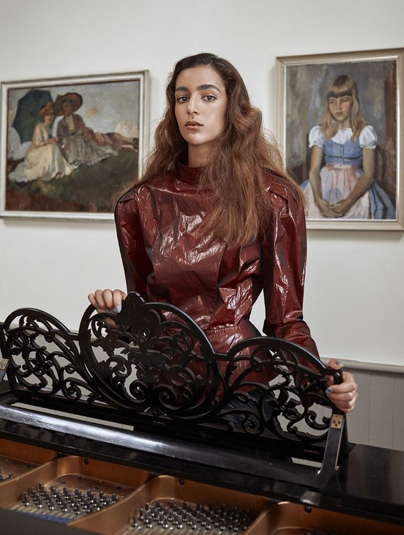 A young woman with wavy brown hair stands by a black piano in front of two framed paintings on a cream-colored wall in an indoor setting.