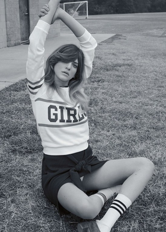 Black and white portrait of a woman in a sweatshirt and skirt sitting cross-legged on a lawn with a soccer net in the background.