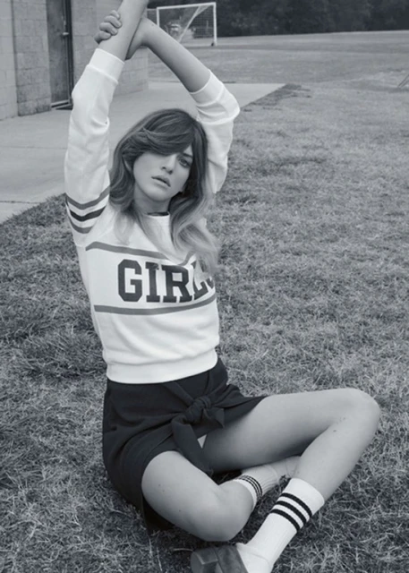 Black and white portrait of a woman in a sweatshirt and skirt sitting cross-legged on a lawn with a soccer net in the background.
