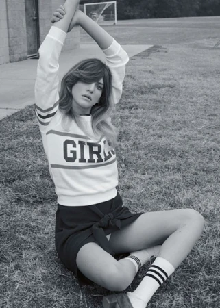 Black and white portrait of a woman in a sweatshirt and skirt sitting cross-legged on a lawn with a soccer net in the background.