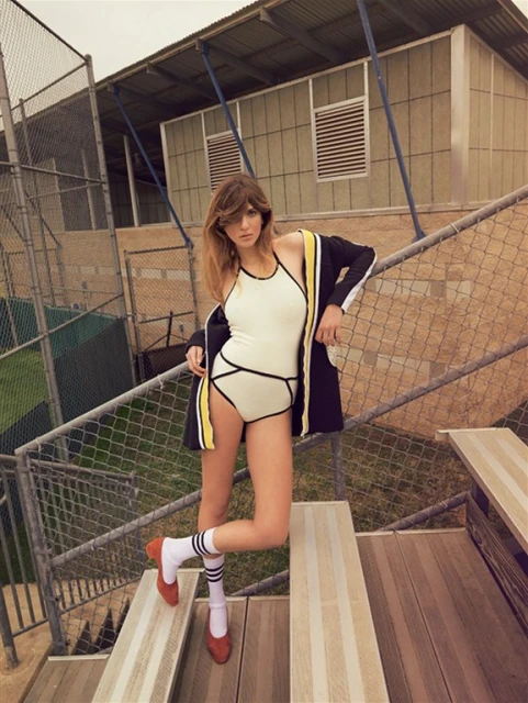A young woman in a white swimsuit with black trim and a jacket stands on wooden bleachers in front of a green sports field.