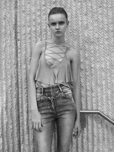 Black and white shot of a young woman in a tank top and jeans, posing against a textured wall with a metal handrail visible.