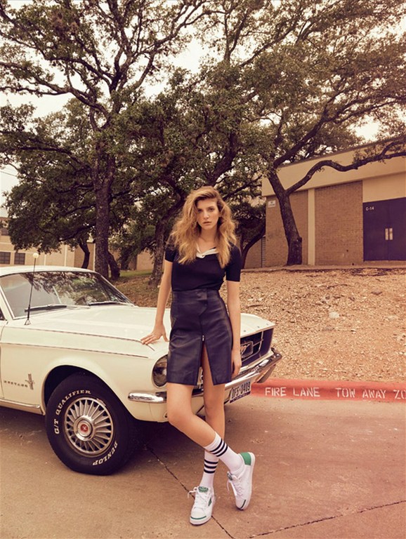 Adult female model poses by a classic car. She wears a black top, leather skirt, white athletic socks, and tennis shoes. The background includes trees and a brick building.