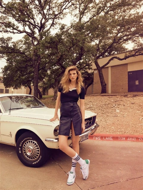 Adult female model poses by a classic car. She wears a black top, leather skirt, white athletic socks, and tennis shoes. The background includes trees and a brick building.