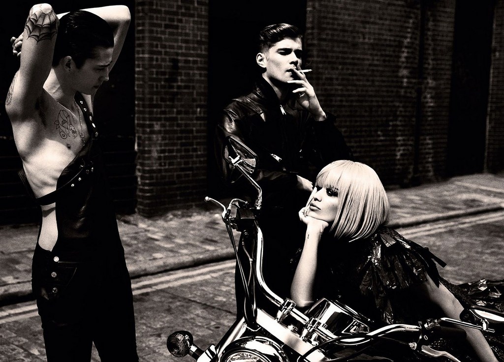 Black and white shot of a woman seated on a motorcycle, with two men standing nearby against a brick building. 