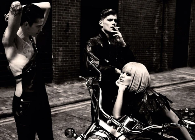 Black and white shot of a woman seated on a motorcycle, with two men standing nearby against a brick building. 