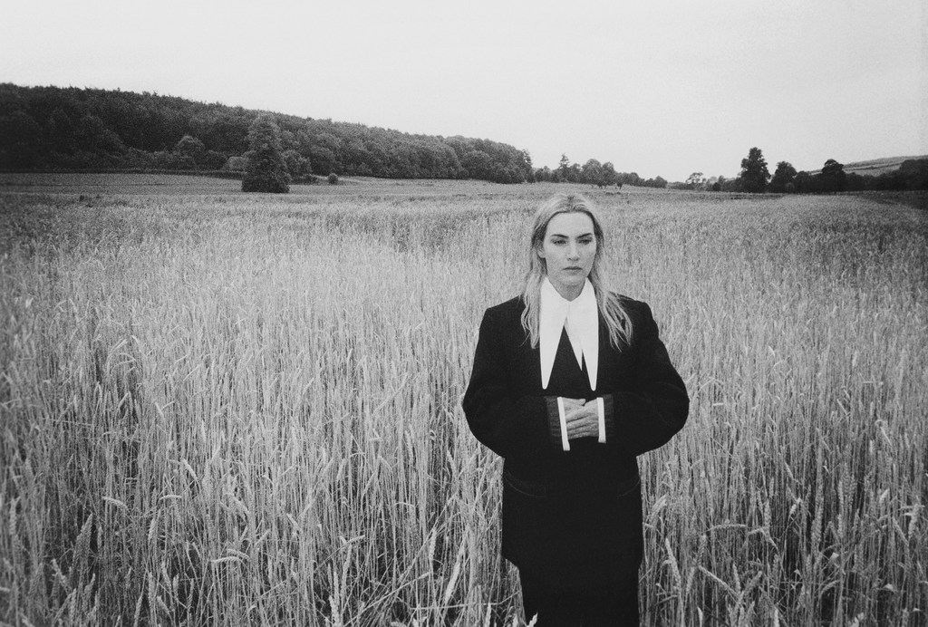 Black and white portrait of a female in a black suit with a white collar standing in a wheat field with a forested horizon.