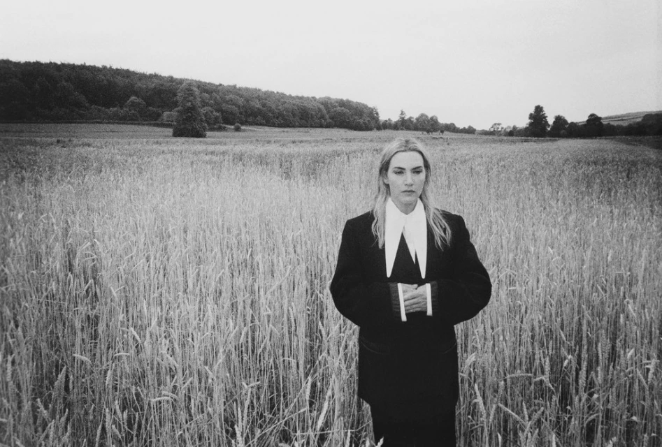 Black and white portrait of a female in a black suit with a white collar standing in a wheat field with a forested horizon.