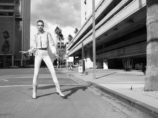 Monochrome fashion shot of a woman in a denim jacket, white jeans, and cowboy boots standing on a city street in a confident pose.