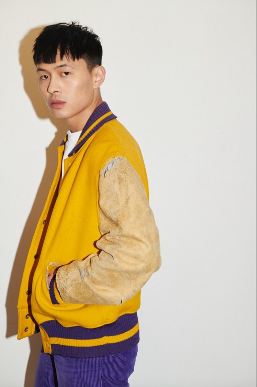 A stylish Asian man poses in a yellow and purple varsity jacket and purple corduroy pants against a light studio backdrop.