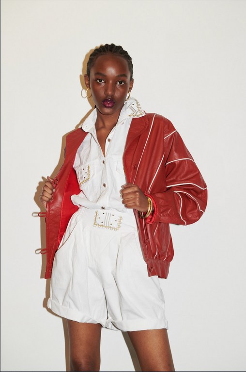 A stylish African woman poses in white shorts and a shirt, complemented by a vibrant red jacket and gold hoops, set against a plain background.