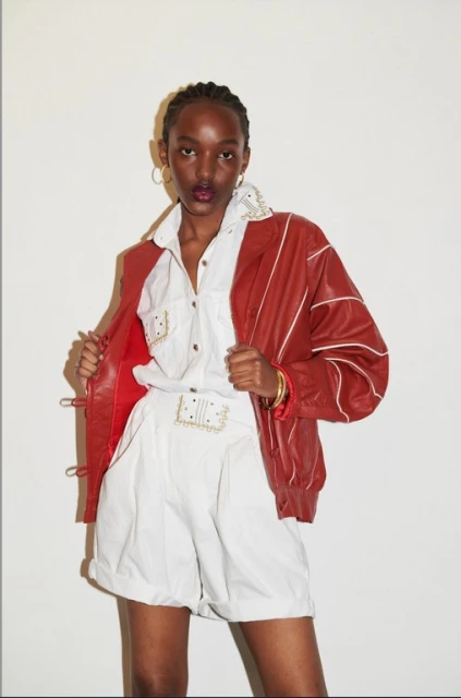 A stylish African woman poses in white shorts and a shirt, complemented by a vibrant red jacket and gold hoops, set against a plain background.