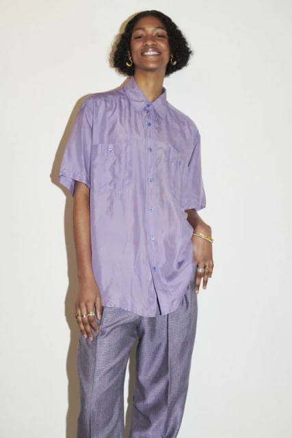 Smiling Black female fashion model in a shiny lavender button-down shirt and patterned trousers stands indoors against a white background.