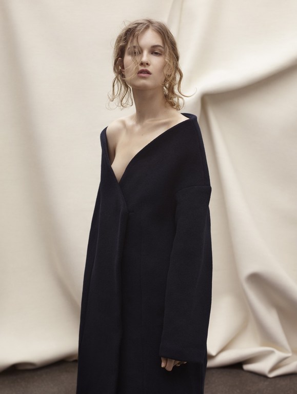 Young woman in a black coat posing against a neutral draped background, fashion studio shot.