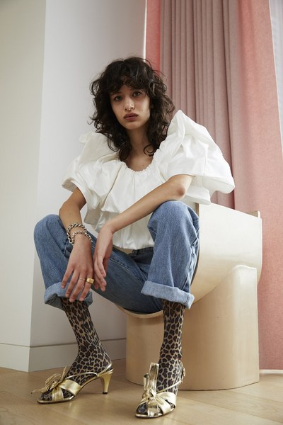 A woman with dark curly hair sits on a sculptural chair wearing a white top, blue jeans, animal-print stockings, and gold high-heeled shoes.
