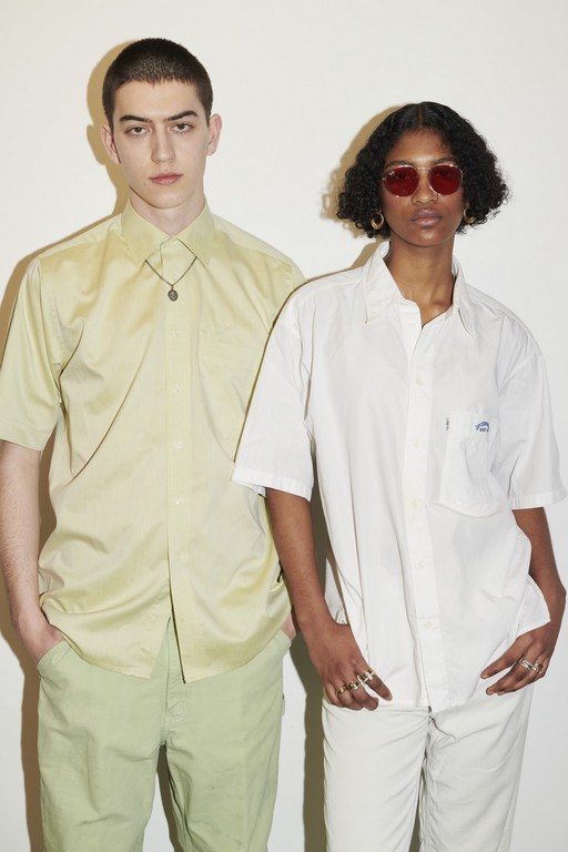 Two models pose against a white wall. A light-skinned man wears a green shirt and pants, and a dark-skinned woman wears a white outfit and red sunglasses.