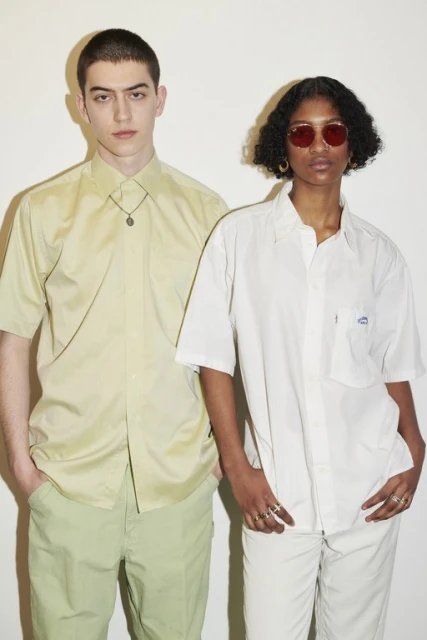 Two models pose against a white wall. A light-skinned man wears a green shirt and pants, and a dark-skinned woman wears a white outfit and red sunglasses.