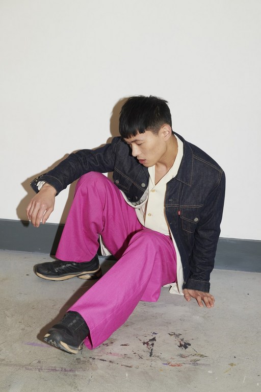 An Asian male model sitting indoors against a plain white wall, wearing a denim jacket, a beige shirt, and bright pink trousers.