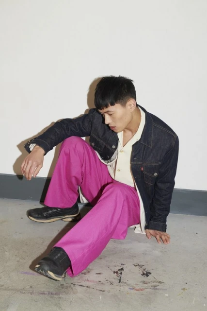 An Asian male model sitting indoors against a plain white wall, wearing a denim jacket, a beige shirt, and bright pink trousers.