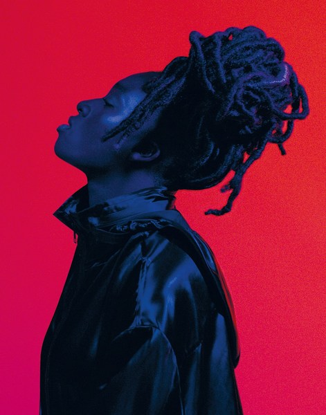 Profile view of a Black woman with dreadlocks against a red and pink gradient background. She is wearing a shiny blue jacket.