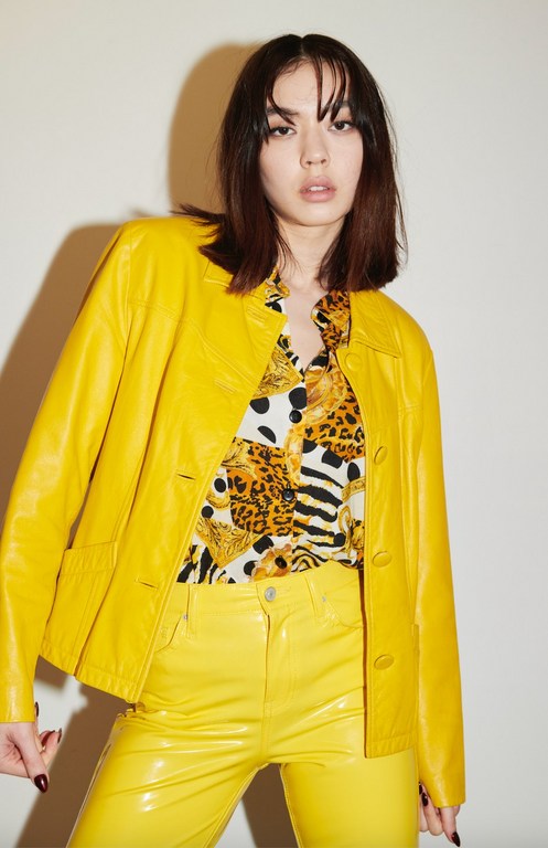 A woman poses in a studio with medium-length brown hair, wearing a bright yellow leather jacket and matching shiny yellow jeans. 