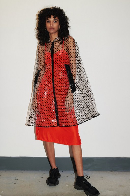 An adult female model poses in a studio wearing a semi-transparent polka dot raincoat over a red dress and chunky black sneakers.