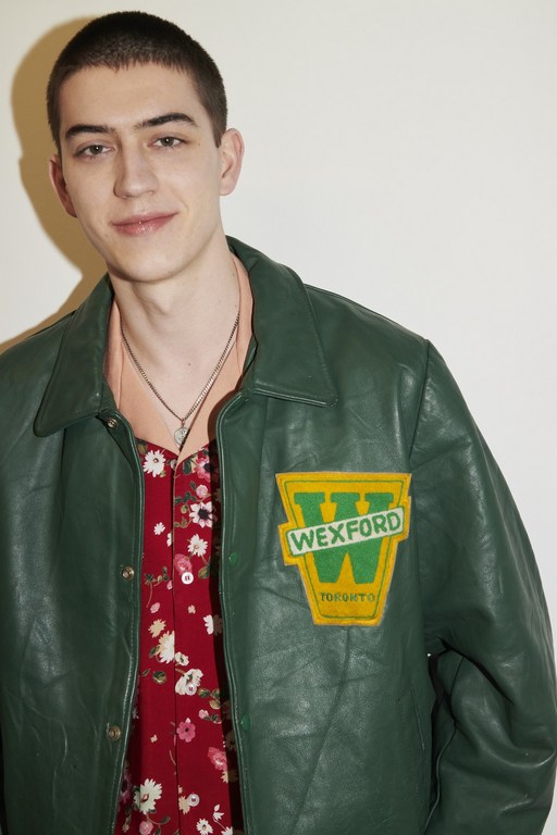 Portrait of a young adult male in a green jacket with a vintage patch over a red floral shirt against a white wall.