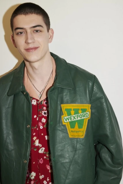 Portrait of a young adult male in a green jacket with a vintage patch over a red floral shirt against a white wall.