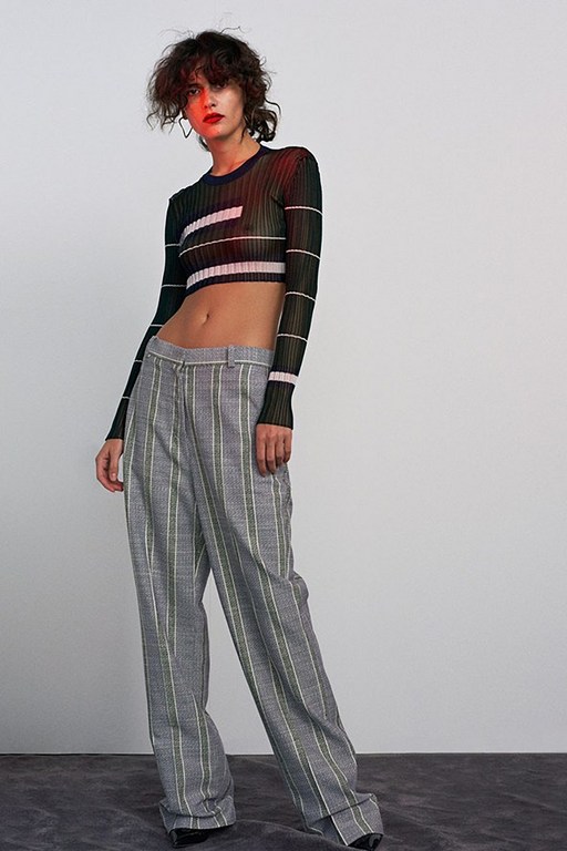 Adult woman in a dark long-sleeved crop top with white stripes and gray, striped wide-leg pants poses against a neutral background.