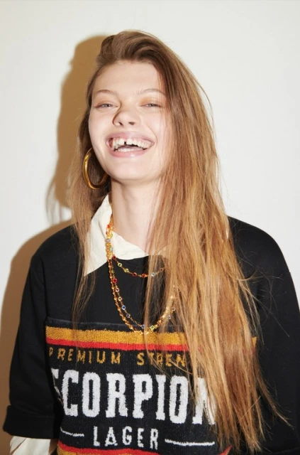 Smiling fair-skinned woman with long brown hair, wearing a black sweatshirt, gold hoop earrings, and beaded necklaces against a white backdrop.