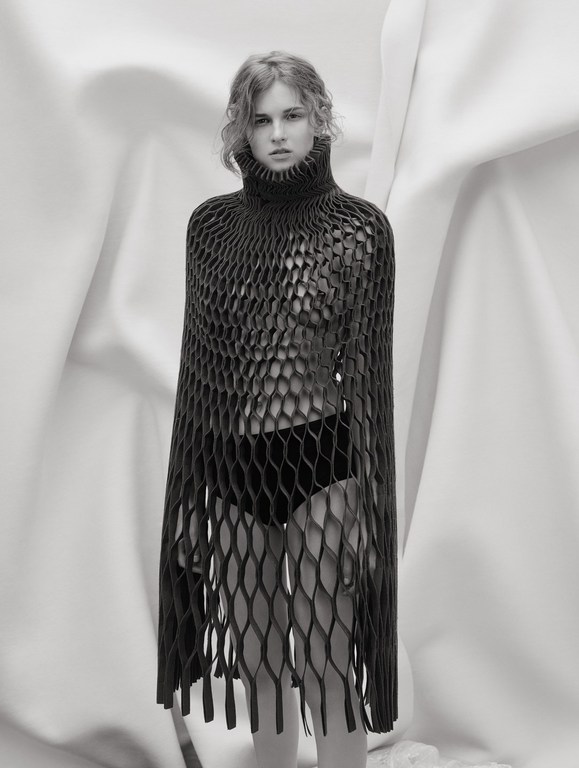 Monochrome portrait of a young woman wearing a black, net-like turtleneck garment, posing in front of draped white fabric backdrop.