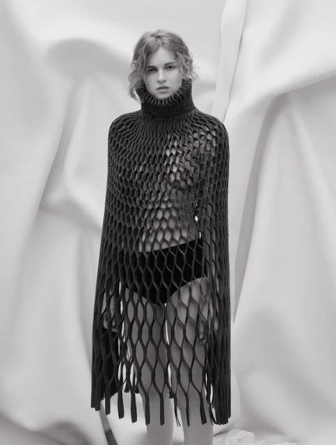 Monochrome portrait of a young woman wearing a black, net-like turtleneck garment, posing in front of draped white fabric backdrop.