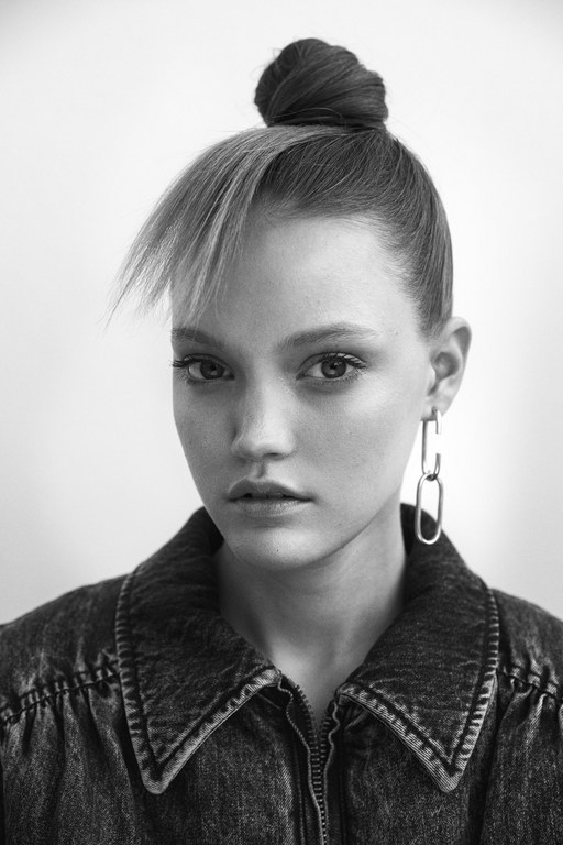 Black and white portrait of an adult woman with her hair in a bun, wearing a denim jacket and silver earrings, against a white backdrop.