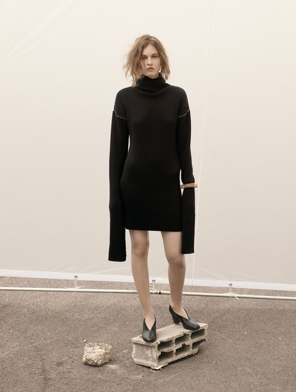 A fair-skinned woman with wavy auburn hair wearing a black turtleneck dress and black heels stands on a concrete block outdoors.