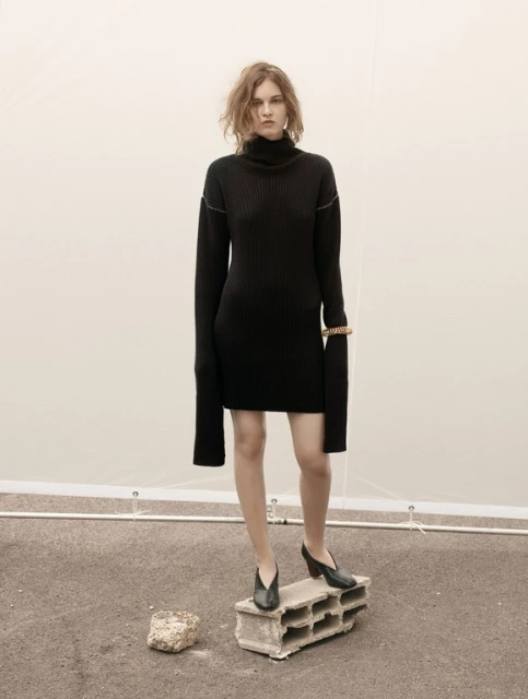 A fair-skinned woman with wavy auburn hair wearing a black turtleneck dress and black heels stands on a concrete block outdoors.
