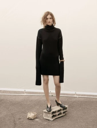 A fair-skinned woman with wavy auburn hair wearing a black turtleneck dress and black heels stands on a concrete block outdoors.