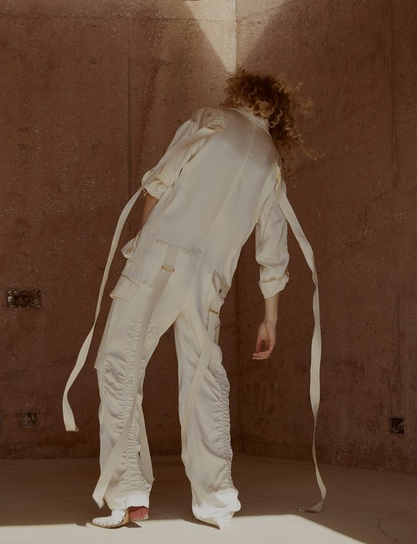Fashion shot of androgynous person in white jumpsuit with trailing ribbons, standing in a room with ochre walls in direct sunlight.