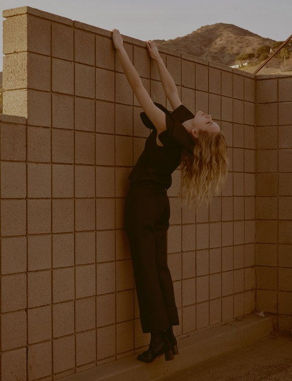 Adult female in black attire stretches against a concrete wall outside, with mountains visible in the background in warm, natural light.