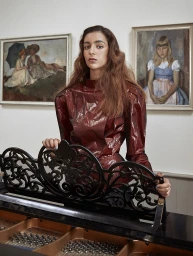 Woman in shiny burgundy blouse posing behind a black upright piano in a gallery with framed landscape and portrait paintings on the wall.
