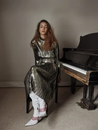 Woman seated on a piano bench next to a grand piano wearing a metallic pleated dress and white boots styled by Natalie Hartley.