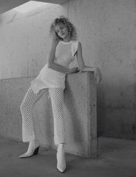 Model with curly blonde hair wearing a netted tunic and trousers leaning against a concrete wall in London