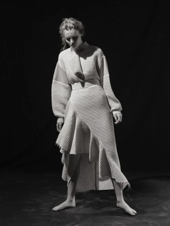 Model standing in a textured cream knit dress with an asymmetric hemline against a dark studio background styled by Natalie Hartley