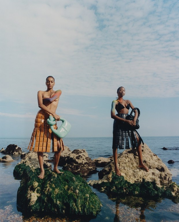 Two women in fashion skirts and bikini tops pose on rocks by the ocean under a partly cloudy sky. Summer fashion editorial.