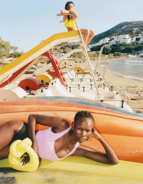 Two fashion models pose on colorful paddle boats on a sunny beach with light blue sky and mountains in the background.
