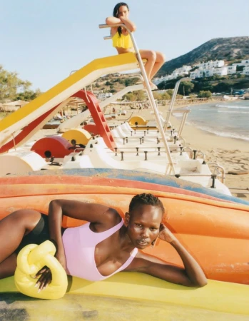 Two fashion models pose on colorful paddle boats on a sunny beach with light blue sky and mountains in the background.